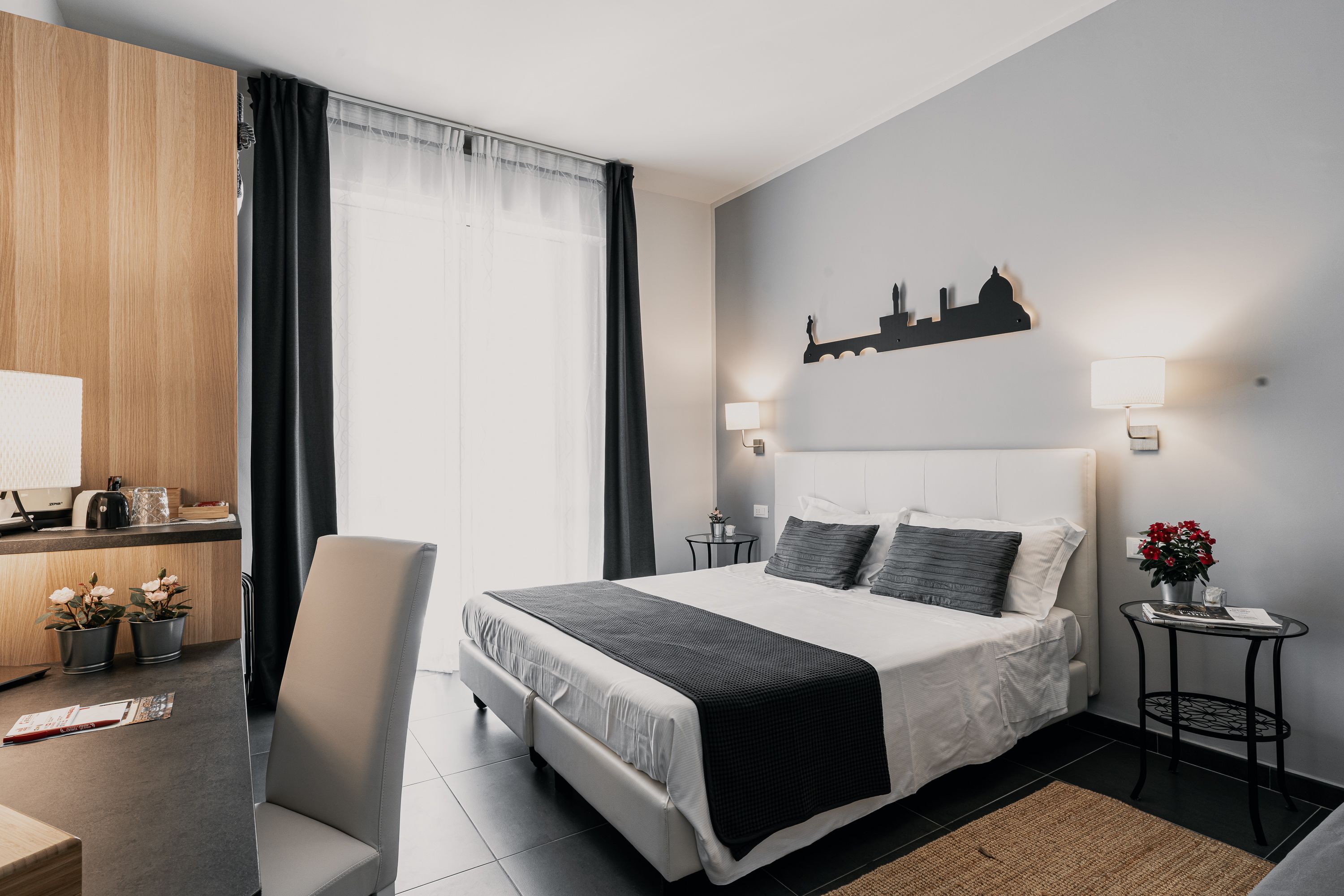 A modern hotel room with a large bed, a desk with a computer, a chair, and a small table with a lamp and flowers.