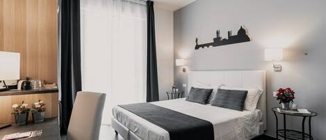 A modern hotel room with a large bed, a desk with a computer, a chair, and a small table with a lamp and flowers.