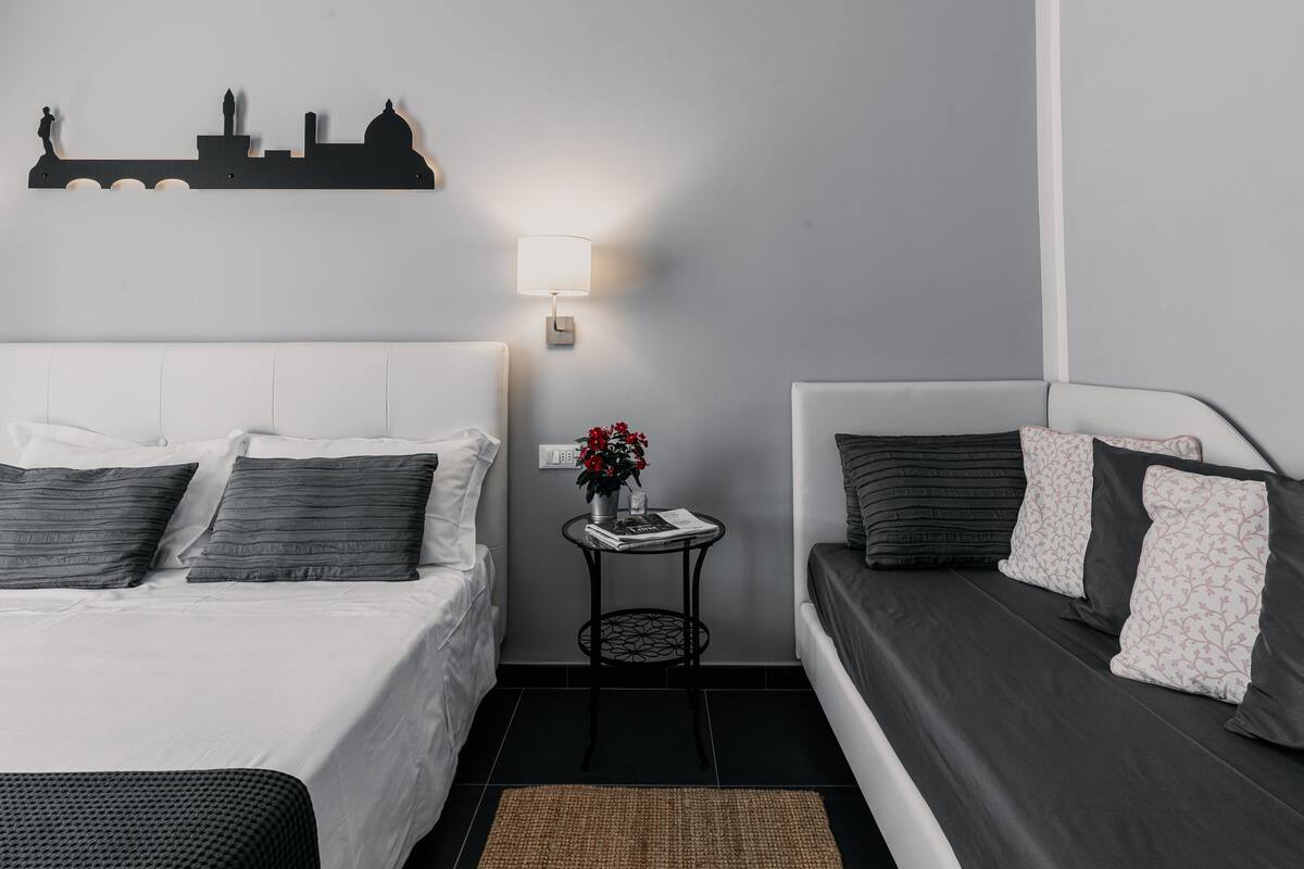 Un dormitorio moderno con una cama grande, un sofá más pequeño, un elemento decorativo en la pared y una pequeña mesa con una lámpara y flores.