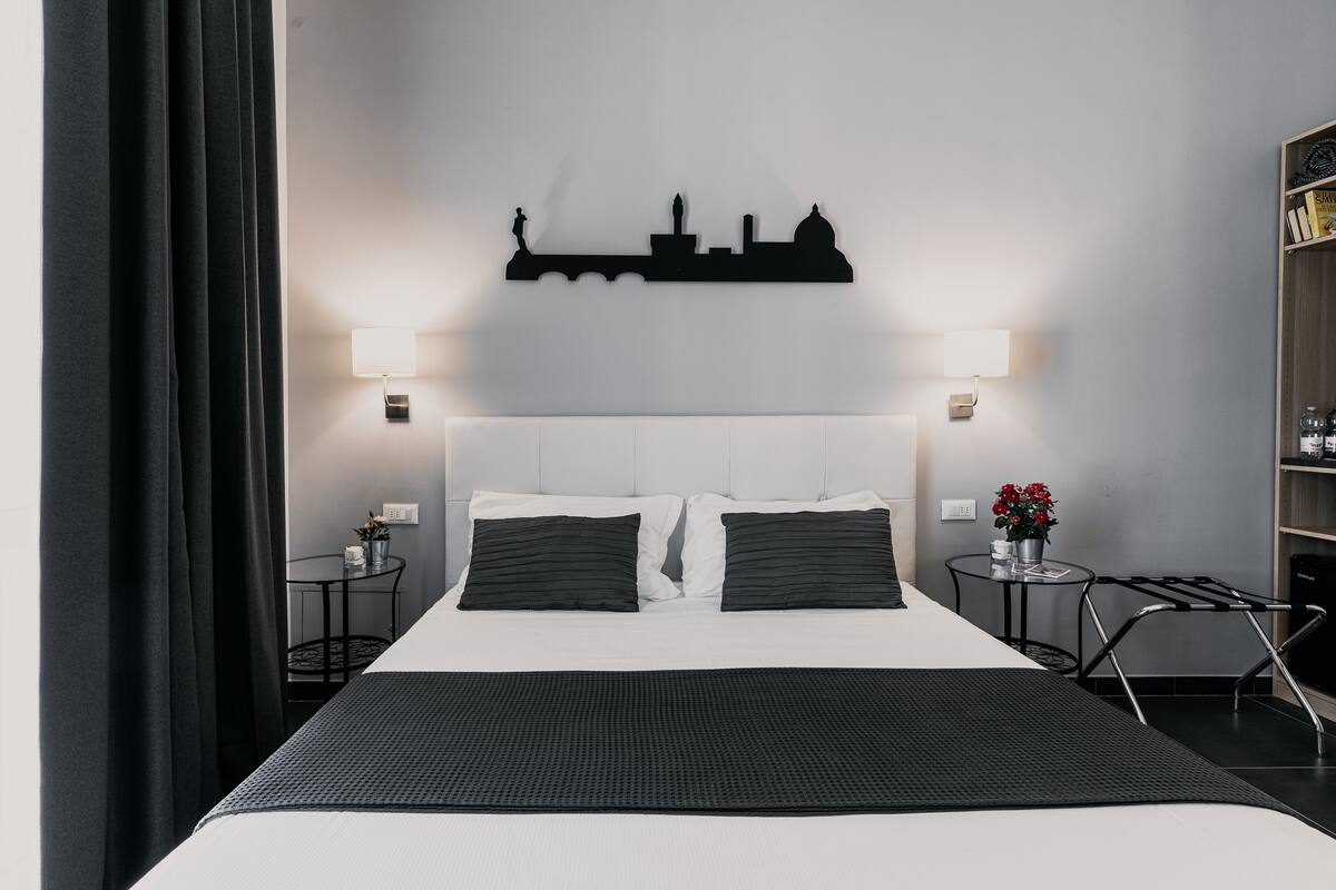 Habitación de hotel con una cama grande, dos mesitas de noche, un cuadro con vista urbana en la pared y una pequeña mesita con una planta.