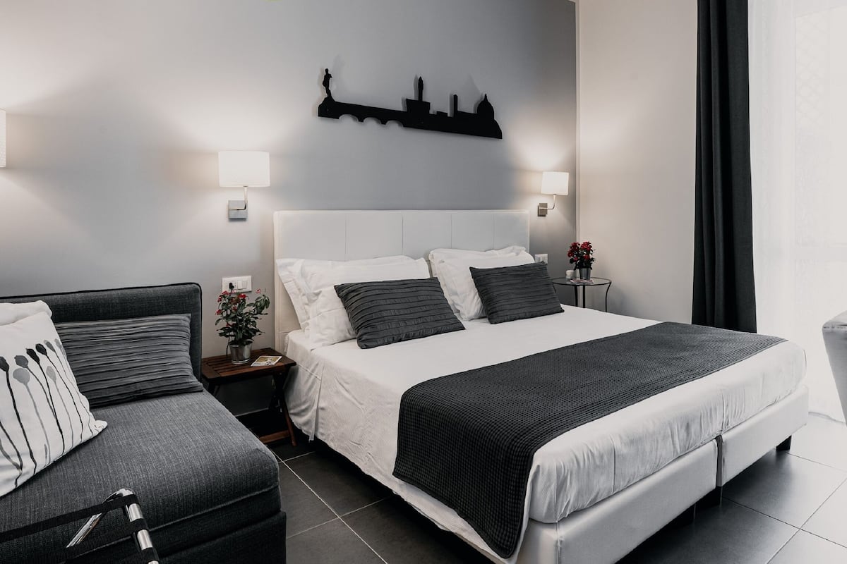 Una habitación de hotel moderna con una cama grande, un sofá más pequeño, una mesita de noche con lámpara y un adorno en la pared.