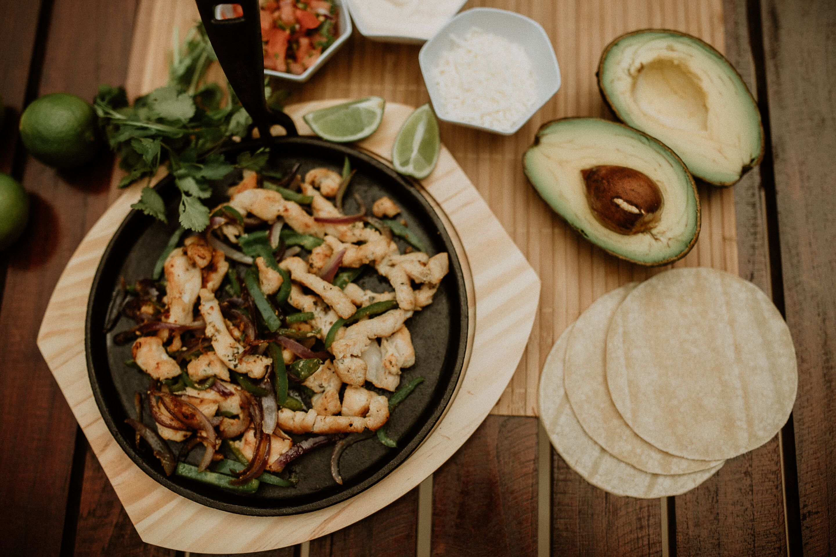 Eine Pfanne mit gegrilltem Hähnchen und Gemüse, Limettenspalten, gewürfeltem Avocado und Tortillas auf einem Holztisch.