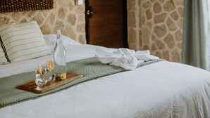 A room with a stone wall, a wooden door, a bed with white bedding, a wicker mirror, and a tray with glasses and orange slices. - Castillo Tambor Resort and Restaurant (Cóbano)