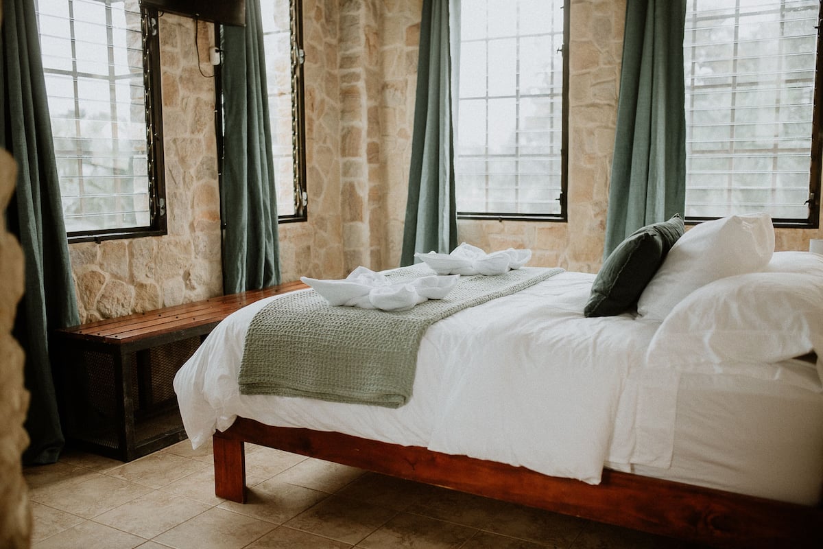 Ein Schlafzimmer mit einem hölzernen Bett, Steinwänden und großen Fenstern mit Vorhängen.