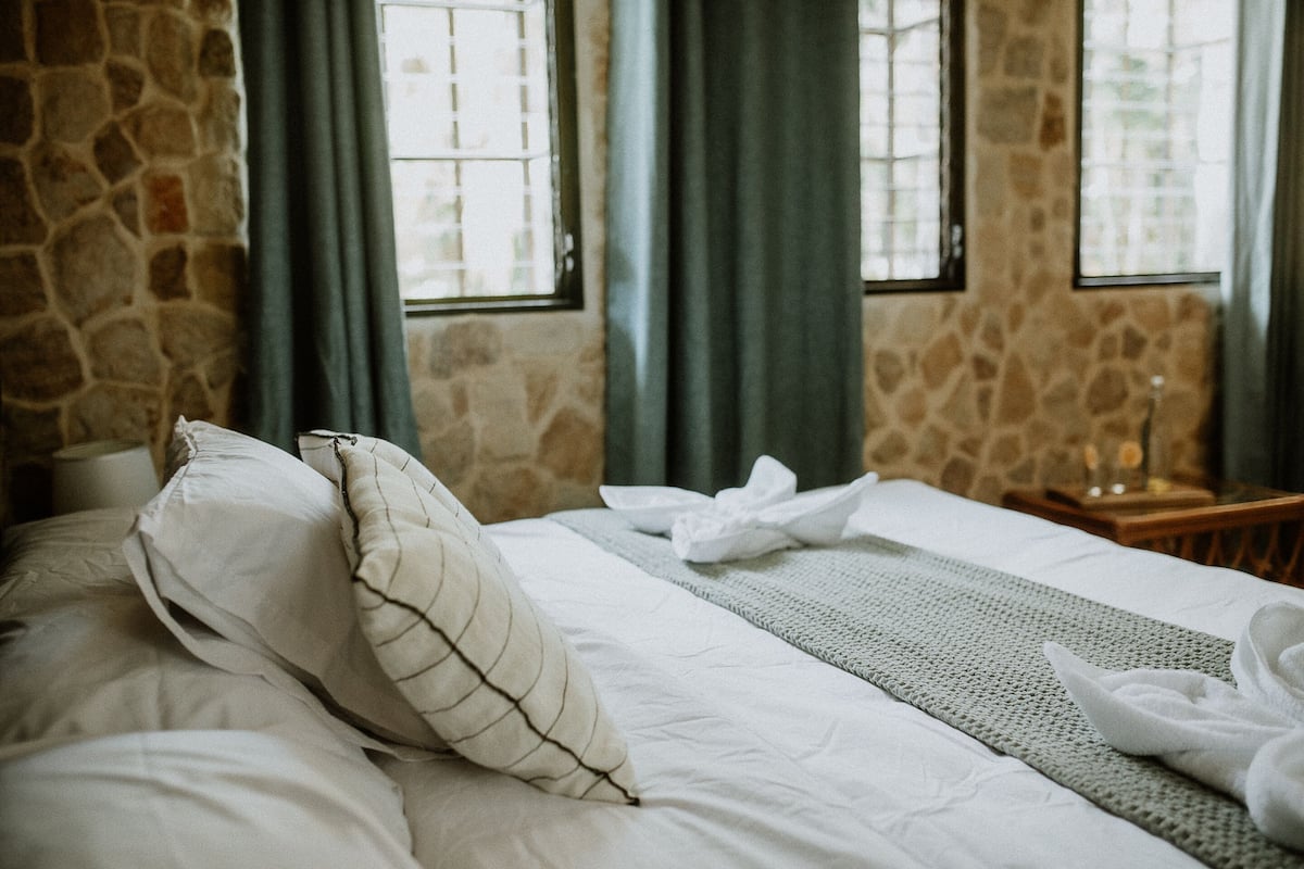 Ein ordentlich bezogenes Bett mit Kissen und Decke, eine Steinwand und ein Fenster mit Vorhängen.