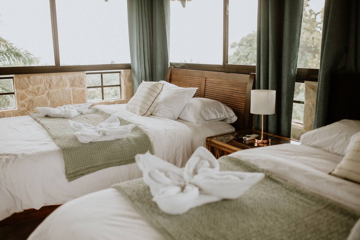 Ein Hotelzimmer mit zwei Betten, einer Steinwand und Blick auf Bäume durch große Fenster.