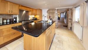 A modern kitchen with wooden cabinets, a large island, and a stone wall. - Camside, Chipping Campden - Taswell Retreats (Chipping Campden)