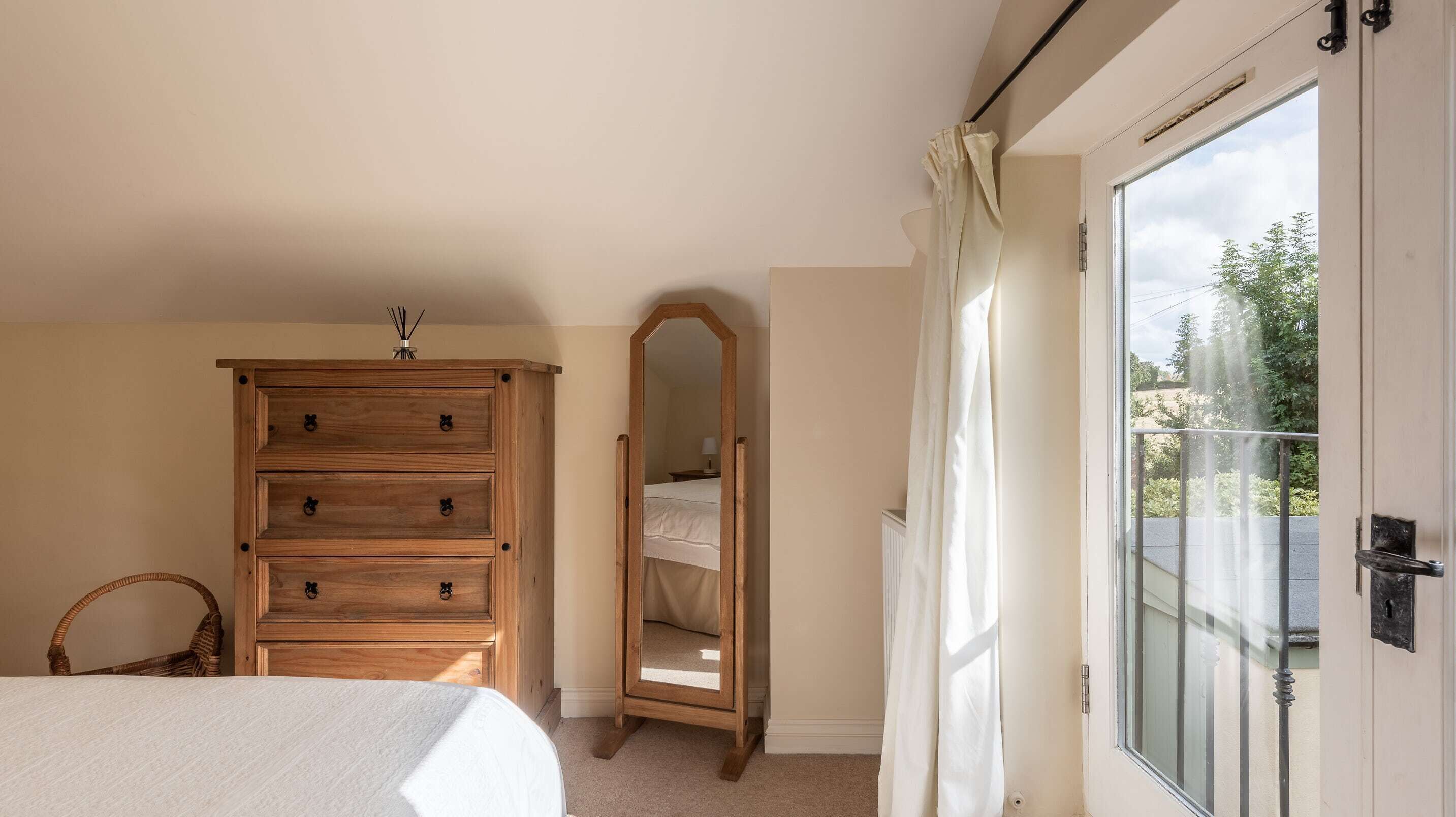 A bedroom with a wooden dresser, a full-length mirror, a bed, and a sliding glass door leading to a balcony.