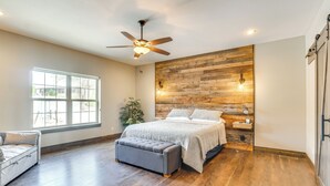 A bedroom with a wooden accent wall, a ceiling fan, a large window, a bed, a bench, and a nightstand with a lamp. - Lavish Retreat w/ Decks, Steps to Table Rock Lake! (Lampe)