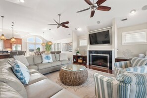 A spacious living room with a large sofa, a coffee table, and a fireplace. There are ceiling fans, pendant lights, and a television mounted on the wall.