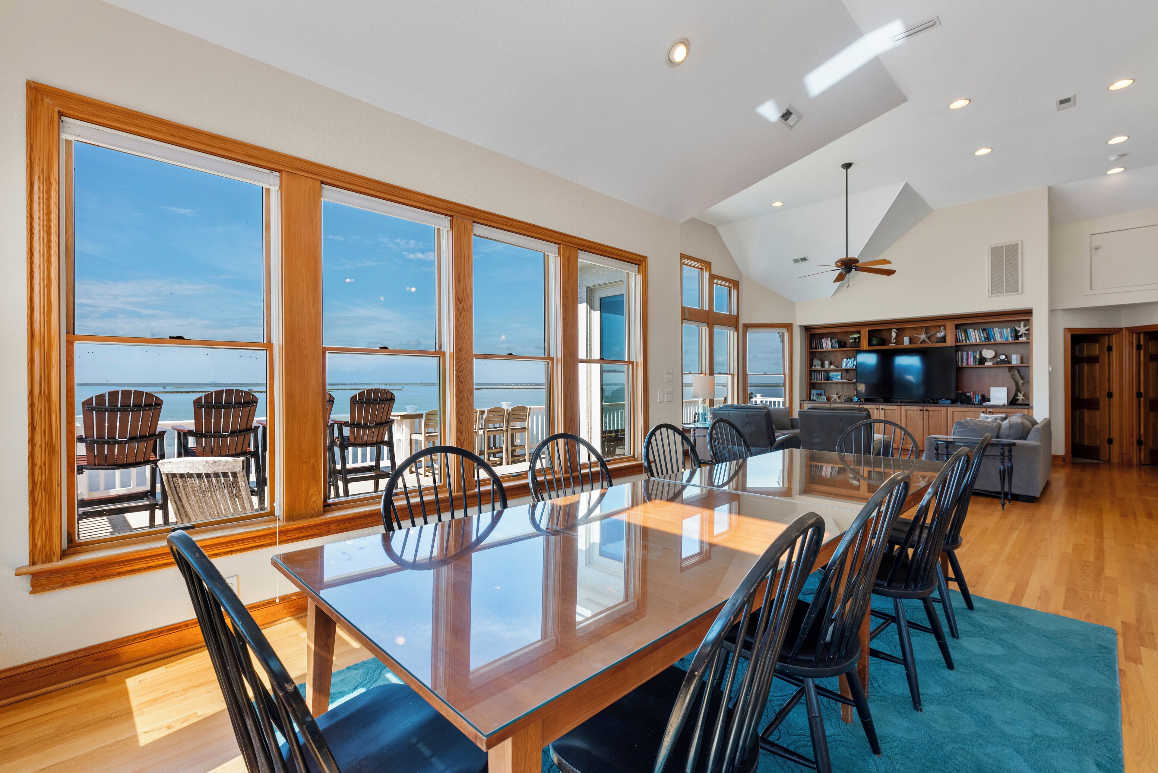 A spacious dining area with a large table and chairs, a kitchen area with a TV and bookshelves, and large windows offering a view of the ocean.
