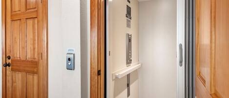 A wooden door with a 'WELCOME' sign, a wall-mounted intercom, and a small elevator with a control panel.
