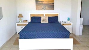 A bedroom with a large bed, two bedside tables, a nightstand, a wall-mounted painting, and a door leading to another room. - Sea Turtle Studio – Upscale Waterview Retreat, Stunning Sunrises & Full Kitchen! (Culebra)