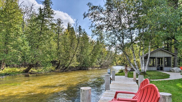 A wooden deck with red Adirondack chairs overlooking a river, surrounded by trees and a building with a glass facade.