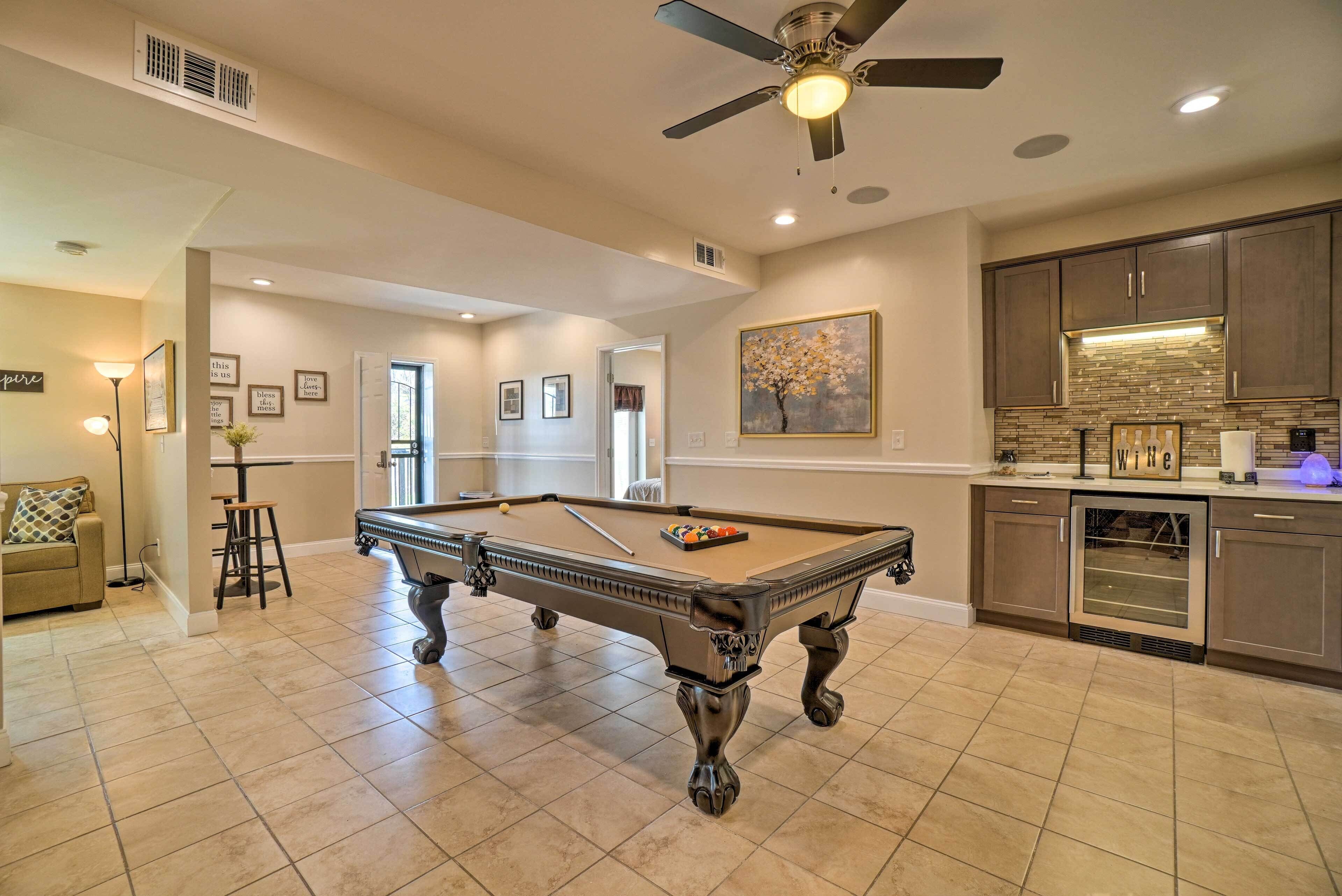 A spacious living area with a pool table, a ceiling fan, and a kitchen area with a wine refrigerator.