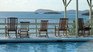 Zona junto a la piscina con sillas de madera y un espacio de estar cubierto con vista al océano.