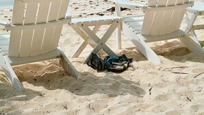 Dos sillas Adirondack blancas sobre una playa arenosa, con un velero al fondo.