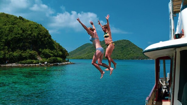 Dos personas saltando desde un bote hacia el agua, con una isla verde al fondo.