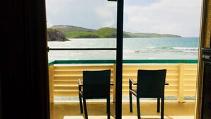Una habitación con un gran ventanal corredera que da a un balcón con dos sillas y una mesa, con vistas a un cuerpo de agua y colinas verdes.