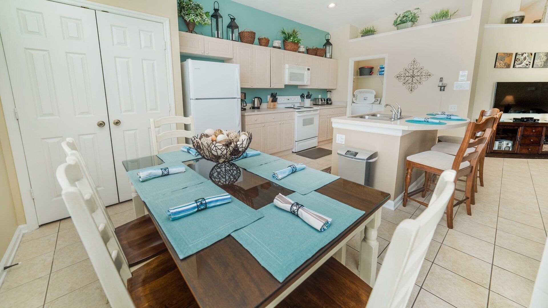 A modern kitchen with white cabinets, a white refrigerator, and a microwave. There is a dining table with blue placemats and a decorative centerpiece.