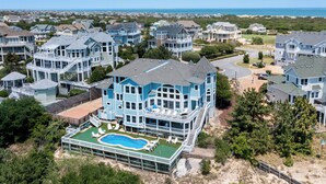 Un'ampia casa celeste circondata da altre case e con una piscina, con una spiaggia sullo sfondo.
