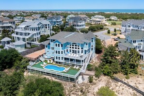 A large, light blue house with a swimming pool, surrounded by other houses and a beach in the background.