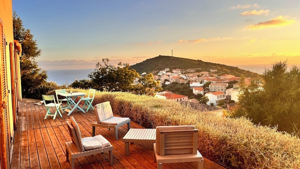 Terraza de madera con mobiliario exterior, con vista a un pueblo costero y colinas al atardecer.