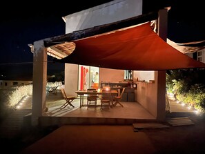 An outdoor dining area with a table and chairs, a kitchen area with a counter and sink, and a large red canopy providing shade.