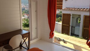 Habitación con un escritorio de madera, una silla blanca, una ventana con cortinas rojas y vistas a edificios y zonas verdes.