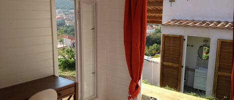 Habitación con un escritorio de madera, una silla blanca, una ventana con cortinas rojas y vistas a edificios y zonas verdes.