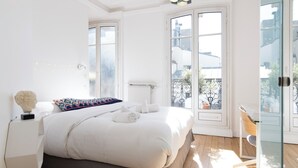 Une chambre lumineuse avec un grand lit, une table de chevet, une lampe et une vue sur le balcon extérieur.