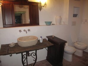Une salle de bain avec un meuble-lavabo en bois, un lavabo rond, un miroir, un luminaire fixé au mur, des toilettes et une étagère ornée d’objets décoratifs.