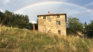 Een rustiek gebouw met een verweerde gevel, omringd door hoog gras en bomen, onder een heldere lucht met een regenboog.