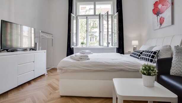 A modern bedroom with a large bed, a TV on a white cabinet, a window with curtains, and a painting of flowers on the wall.