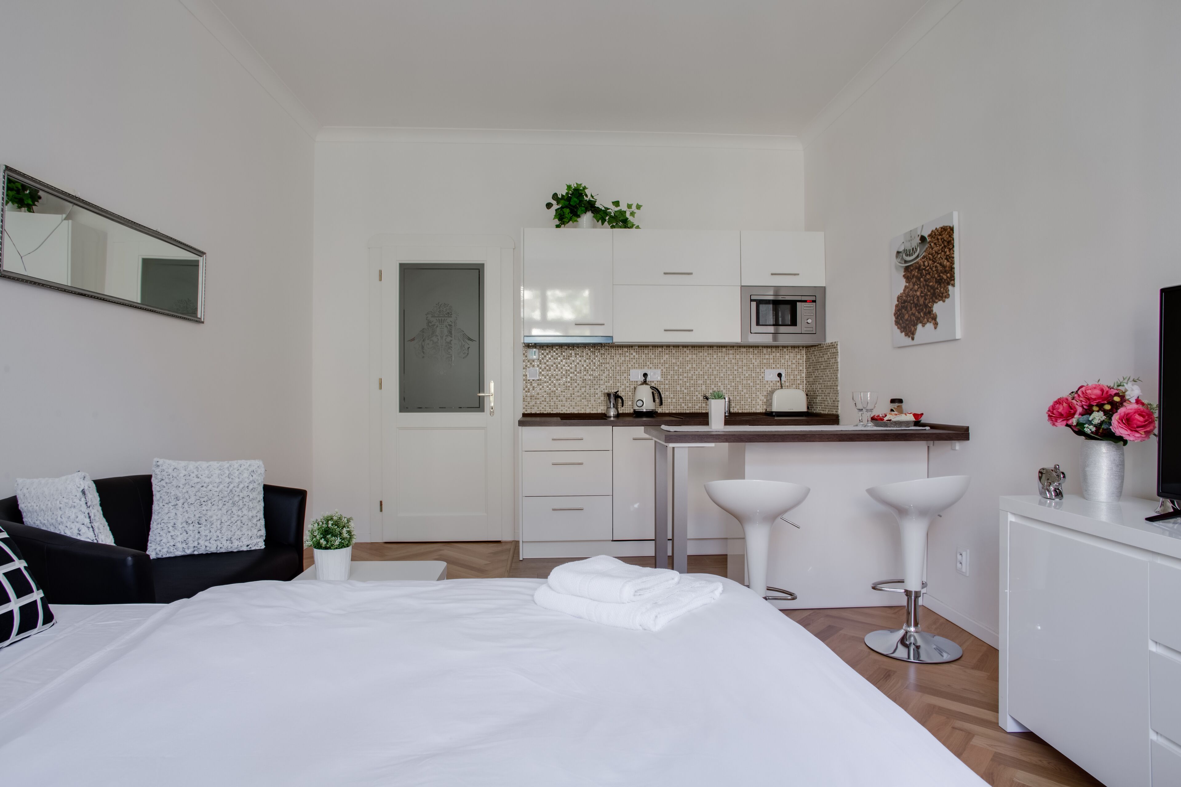 A modern hotel room with a large bed, a black leather sofa, a small table, a kitchen area with a microwave, and a bar with two stools.