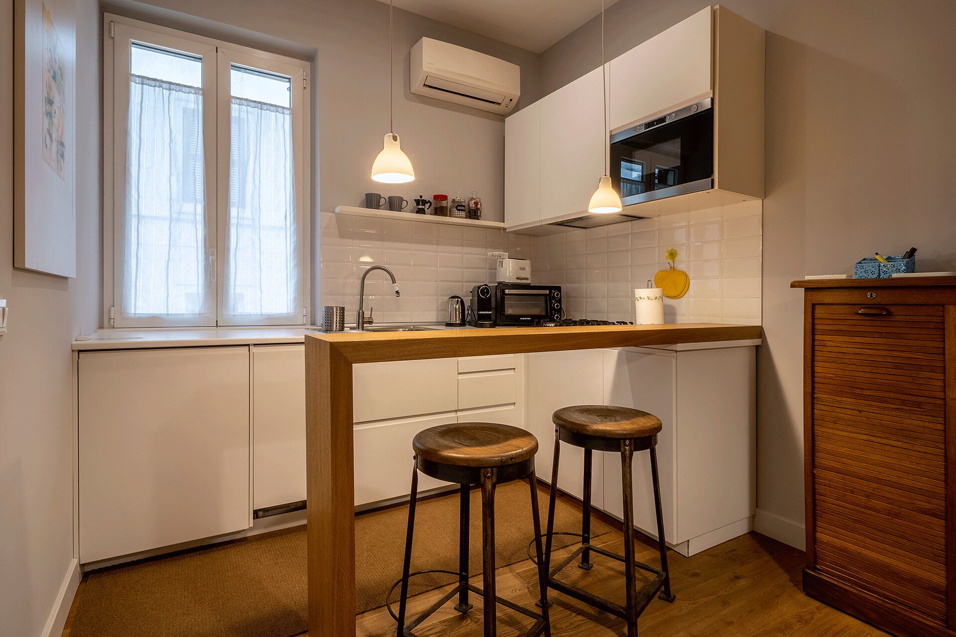 Une cuisine compacte avec des meubles blancs, un plan de travail en bois, deux tabourets, un four à micro-ondes et un appareil de climatisation.