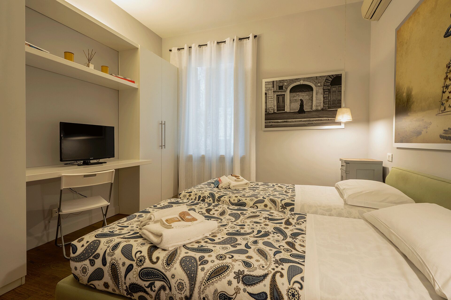 Ein Hotelzimmer mit einem Bett, einem Schreibtisch mit Fernseher, einem Sessel, einem Fenster mit Vorhängen und einem Bild an der Wand.