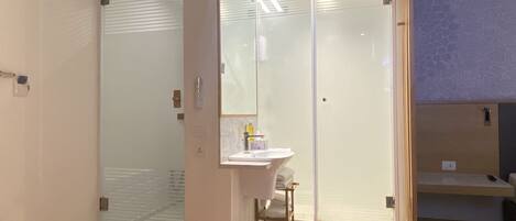 Habitación de hotel con baño que incluye cabina de ducha con mampara de vidrio, lavabo con artículos de aseo y un estante pequeño.