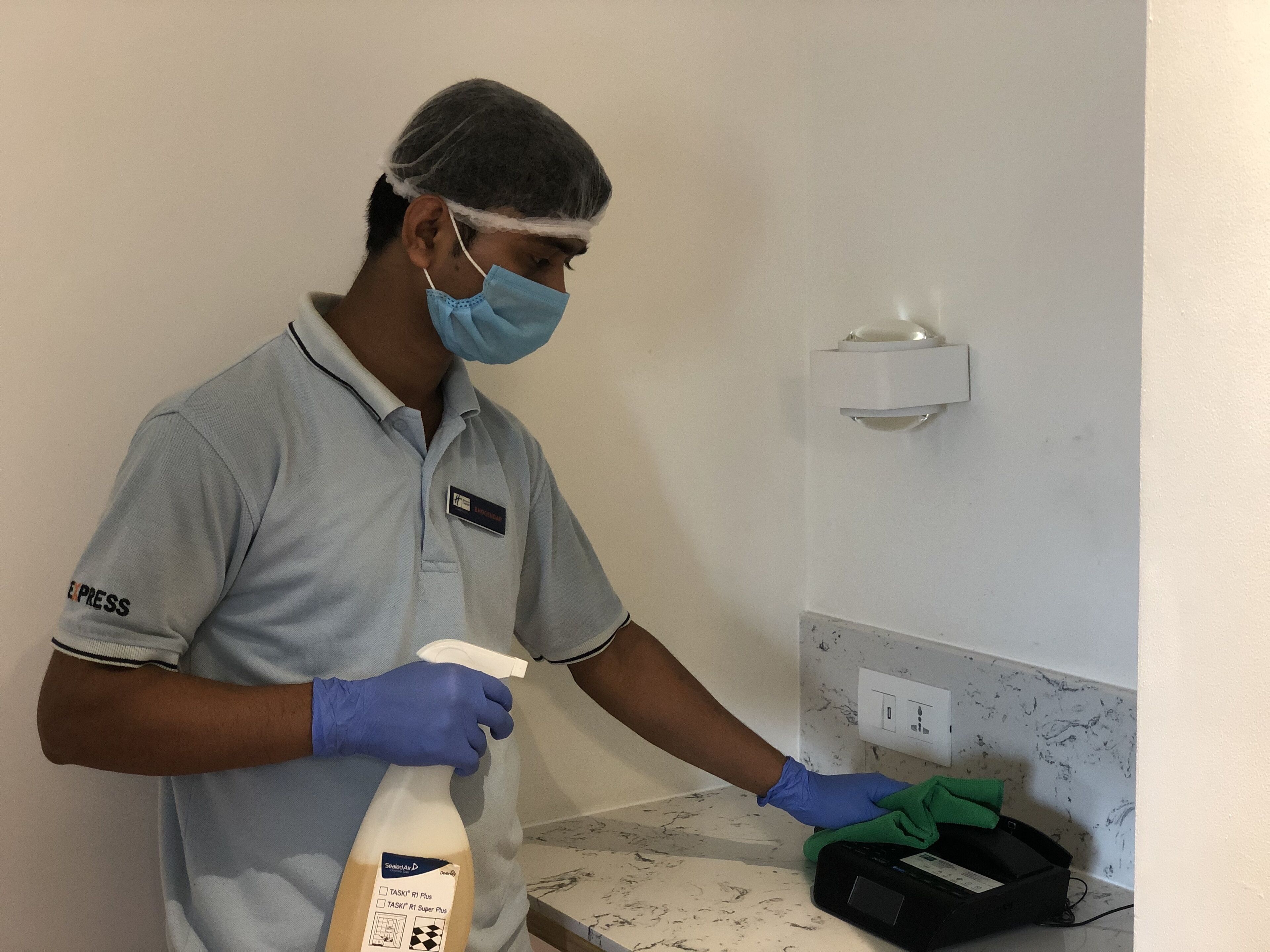 Een persoon in lichtblauw uniform, met naambadje, een haarnet en handschoenen, die een sprayfles en een doek vasthoudt, terwijl een marmeren aanrecht wordt schoongemaakt in een kamer met een witte muur en lichtschakelaar.