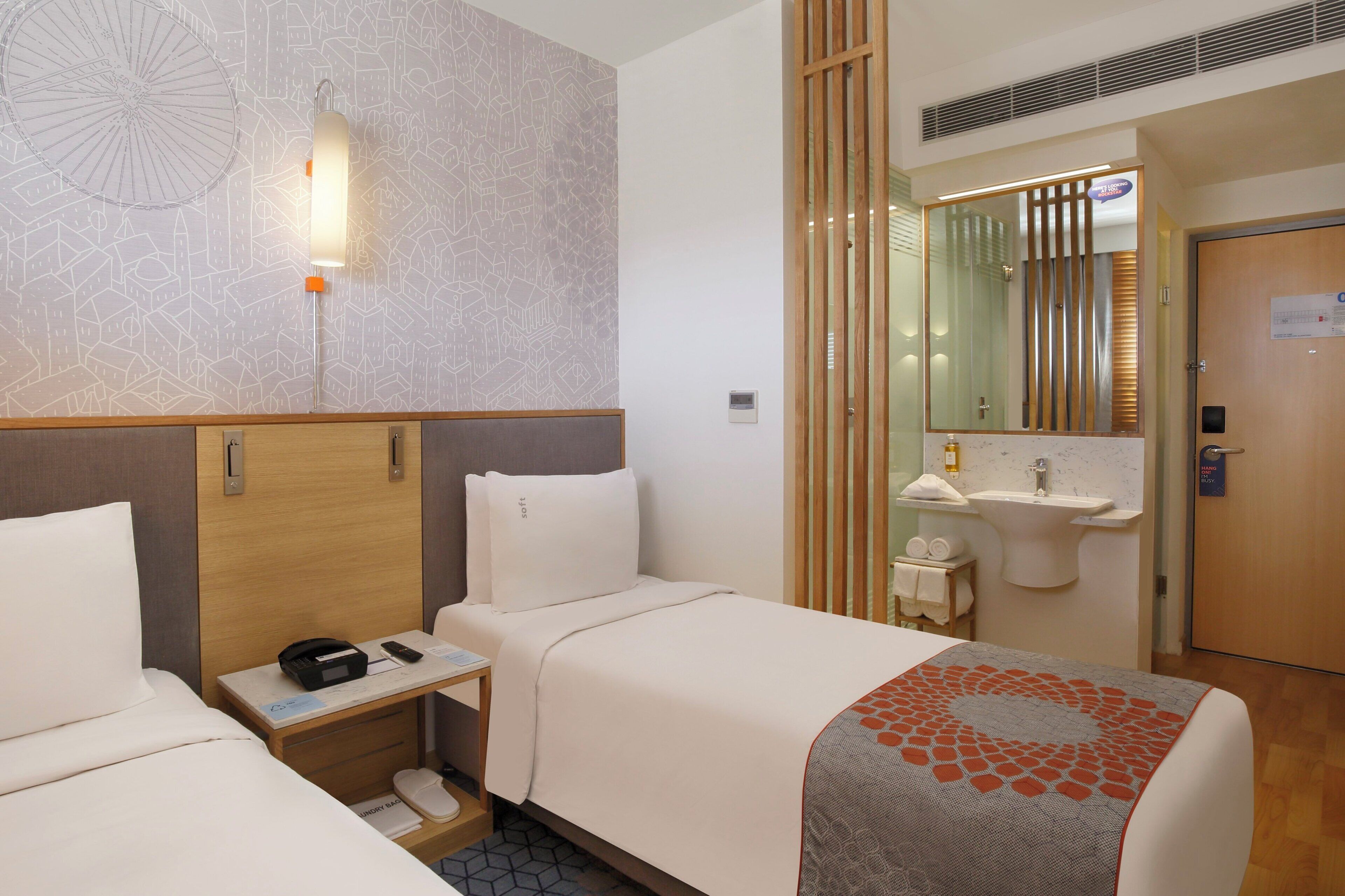 A hotel room with two beds, a bedside table with a phone and a charger, a small bathroom with a sink and mirror, and a decorative wall pattern.
