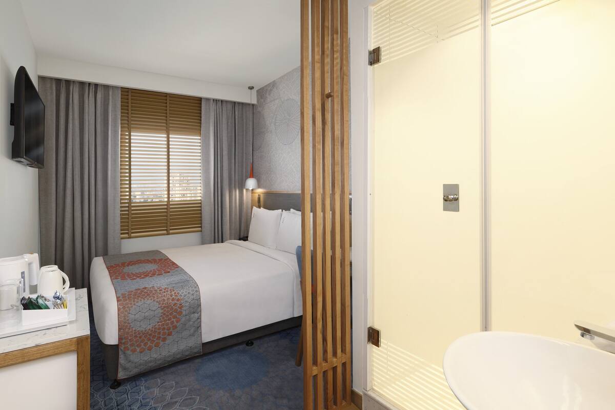 Une chambre d’hôtel comprenant un lit, une table de chevet avec une machine à café, une fenêtre avec des stores, une cloison en bois et une salle de bain avec un lavabo et un miroir.