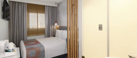 A hotel room with a bed, bedside table with a coffee maker, a window with blinds, a wooden partition, and a bathroom with a sink and mirror.