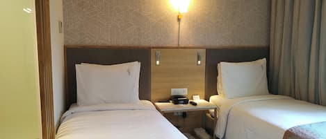 Habitación de hotel con dos camas, una mesita de noche con un teléfono y un reloj digital, y una lámpara fijada a la pared.
