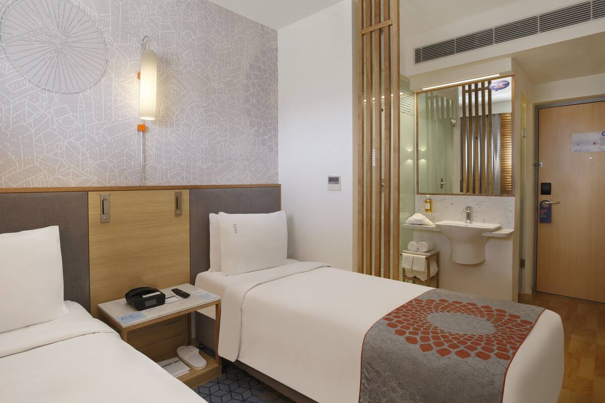Une chambre d’hôtel avec deux lits, une table de chevet avec un téléphone et un chargeur, un petit bureau avec une chaise, un miroir, un lavabo et une porte.