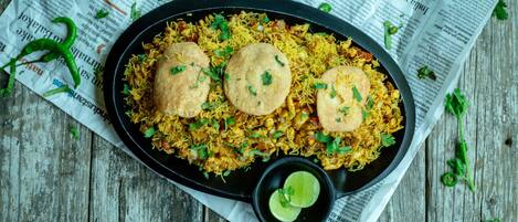 A plate of biryani with two puris on top, garnished with herbs, and a small bowl of lime wedges.