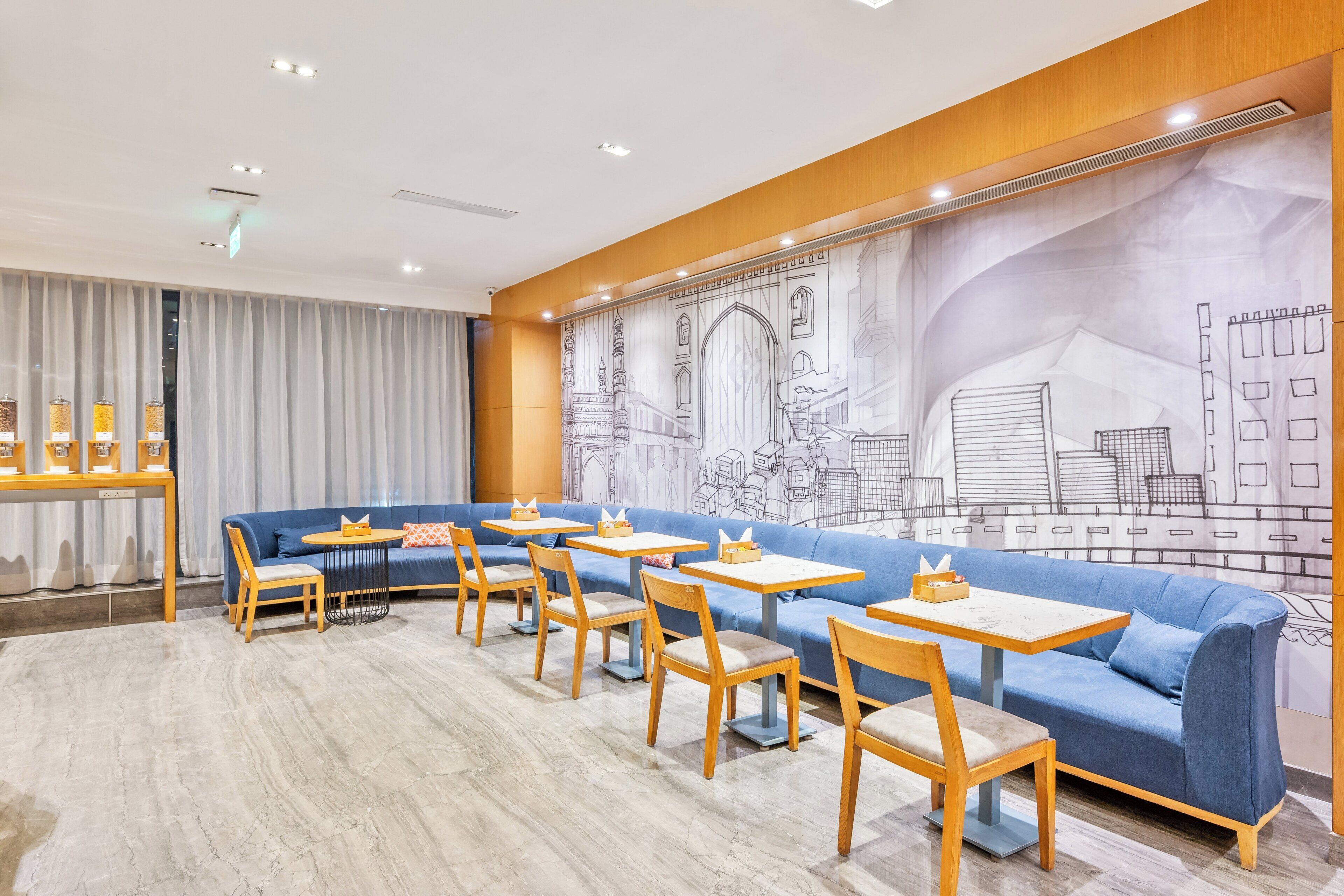 A modern cafe with blue sofas, wooden tables, and a large mural of architectural drawings on the wall.