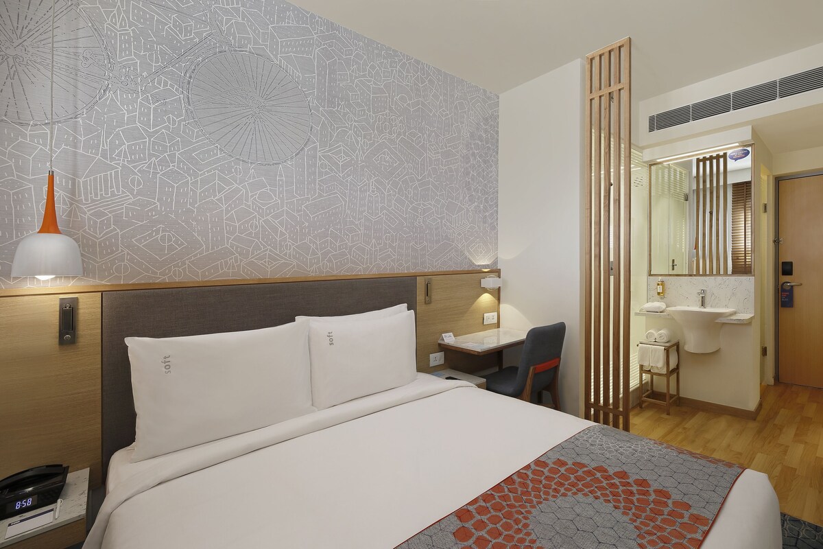 Een hotelkamer met een bed, nachtkastje, lamp en een badkamer met wastafel, spiegel en handdoekhouder.