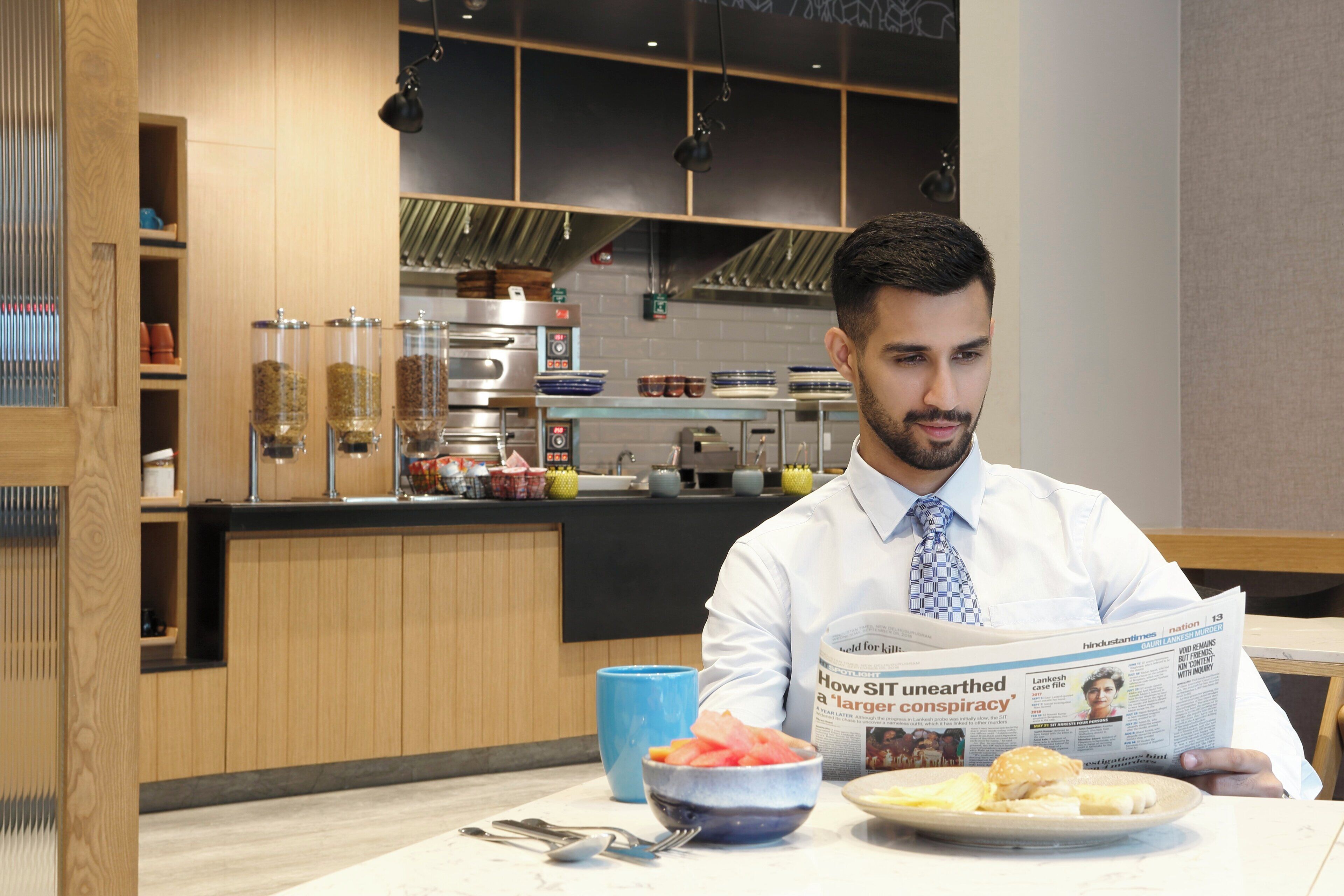 A person reading a newspaper at a table with a bowl of fruit and a plate of food.