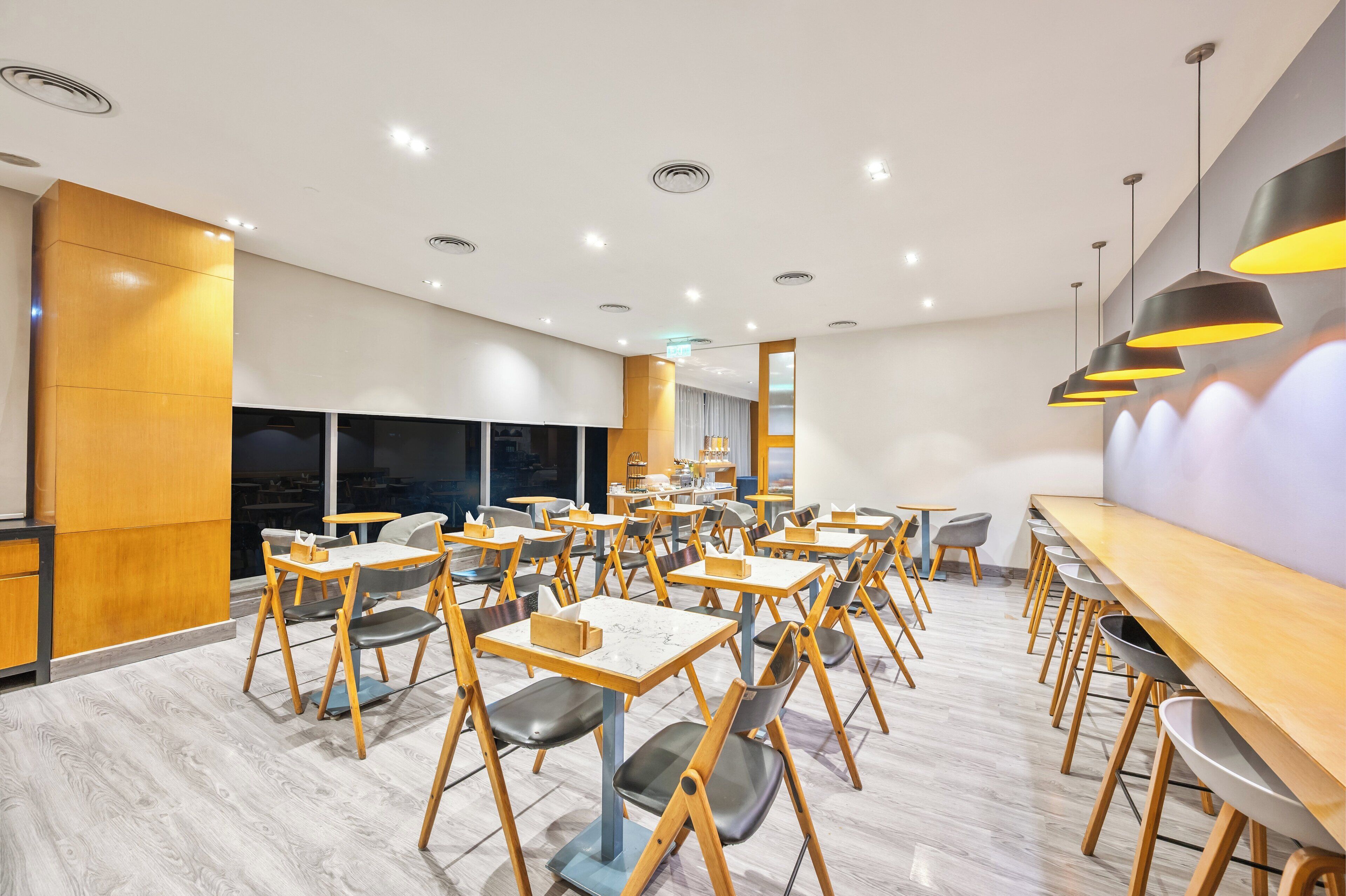 A modern dining area with wooden tables and chairs, large windows, and recessed ceiling lights.