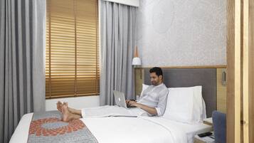 A hotel room with a bed, a laptop, a bedside table, and a window with blinds.